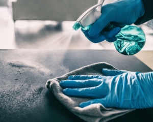 Bilde av person med blå hansker som sprayer desinfeksjonsmiddel på en bordflate og tørker med mikrofiberklut.